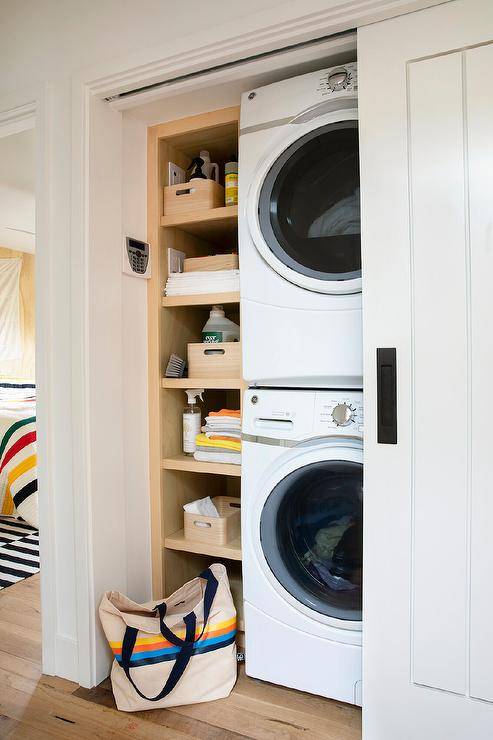 A hallway closet laundry room boasts a white sliding door with oil rubbed bronze hardware and a stacked white front loading washer and dryer fixed beside built-in oak shelves.