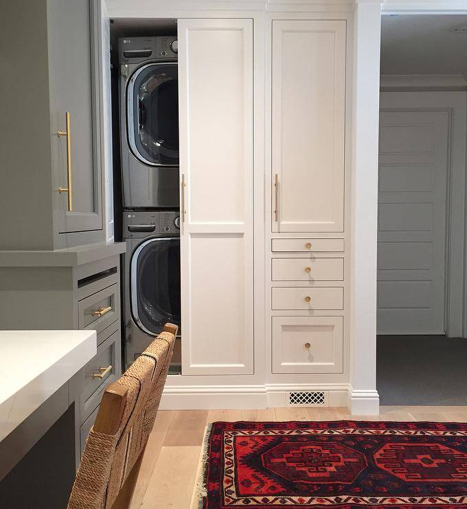 Laundry room features hidden stacked washer and dryer concealed behind bi-fold cabinet doors across from a gray built-in desk flanked by tall gray built-in cabinets adorned with long brass pulls.