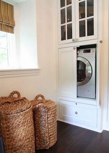 Dream walk-in closet features washer dryer combo hidden within built-in white cabinets with glass front uppers, accented with mercury glass pulls alongside a pair of seagrass laundry hampers.