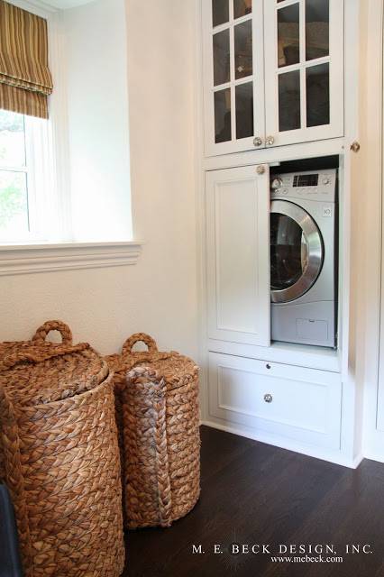 Dream walk-in closet features washer dryer combo hidden within built-in white cabinets with glass front uppers, accented with mercury glass pulls alongside a pair of seagrass laundry hampers.