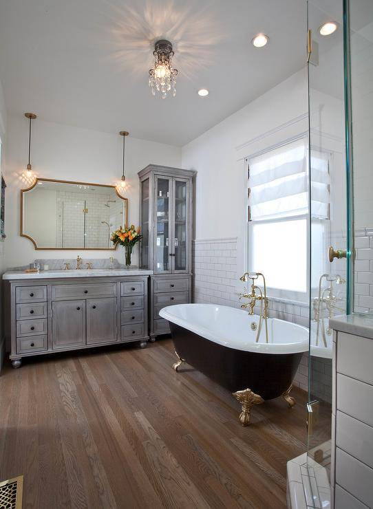 Elegant bathroom features an eye-catching black and gold clawfoot bathtub paired with a polished brass vintage tub filler and positioned beneath a window dressed in a white roman shade. The wall behind the tub is half clad in white subway backsplash tiles illuminated by recessed lighting a small crystal chandelier.