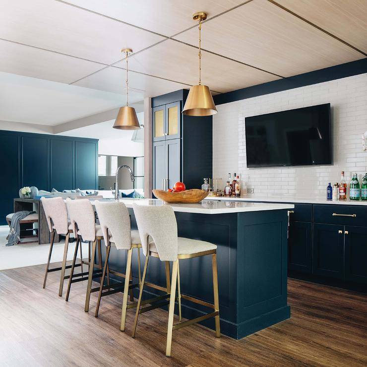 Sophisticated basement wet bar features gray and gold barstools that compliment a blue shaker wet bar island boasting white quartz countertop, facing a wall with white glass brick pattern tiles with mounted tv, flanked by dark blue shaker wet bar cabinets accented with brass hardware. The room is lit by brass lanterns from above.