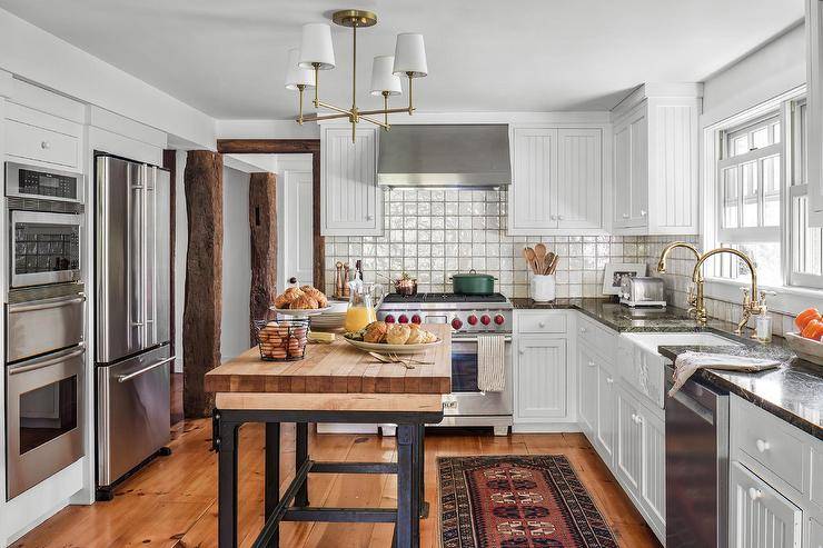 Spacious cottage kitchen features a freestanding wood and iron island lit by a brass chandelier, a vintage runner, white cabinets and stainless steel refrigerator and oven.