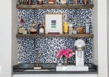 Black and white leopard print wallpaper accents a wet bar boasting brown wood floating shelves mounted above an antique brass faucet fitted to a black countertop accenting white cabinets.