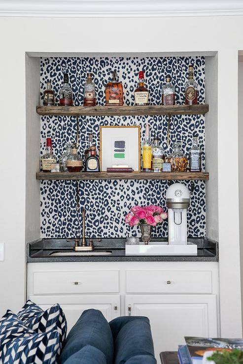 Black and white leopard print wallpaper accents a wet bar boasting brown wood floating shelves mounted above an antique brass faucet fitted to a black countertop accenting white cabinets.