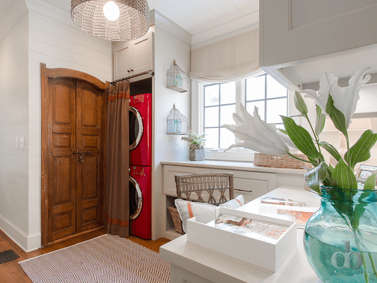 Cottage laundry room features stacked red washer and dryer enclosed with gray cabinets hidden behind brown and red curtains next to a closet fitted with wood double doors. Laundry room with work space boasts a desk situated under cabinets atop a chevron rug next to a laundry folding station under window.