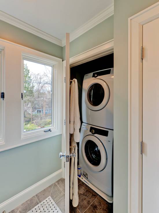 Neat cupboard off bathroom with stackable white front loading washer and dryer. Inside the cupboard door hang towel rails with fresh white towels. The bathroom walls are painted a sage green which pairs with tiled floors with a marble basketweave tiled rug.
