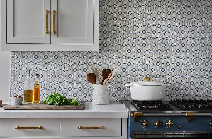White kitchen cabinets donning antique brass hardware are mounted to blue and gray mosaic backsplash tiles complementing a blue French stove.