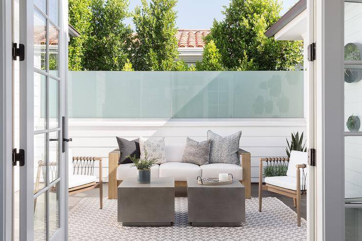 French doors open to a patio boasting concrete cube accent tables placed on a gray diamond pattern rug covering gray tiles. Teak and rope chairs flank a teak sofa positioned in front of a shiplap and frosted glass wall.