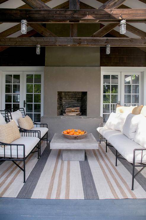 Gray and tan striped outdoor rug with a concrete table centered between wrought iron sofa and chairs. A truss patio ceiling displays nautical industrial styled lights over a well-designed patio display.