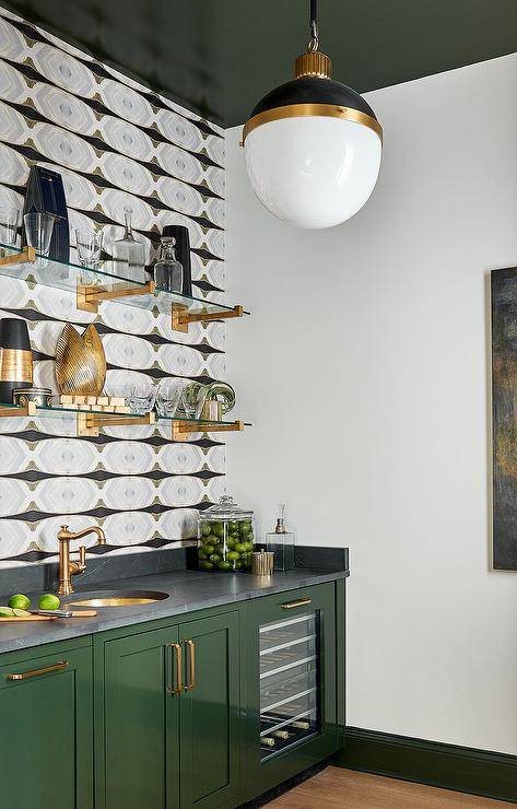 Stylish contemporary wet bar boasts glossy green lacquered cabinets donning brass pulls and a soapstone countertop located over a glass front wine cooler and finished with a round brass sink with a brass hook and spout faucet. Brass and glass shelves are mounted over the sink from a wall covered in black and gray wallpaper, a black and gold Hicks Pendant hangs from a glossy green painted ceiling.