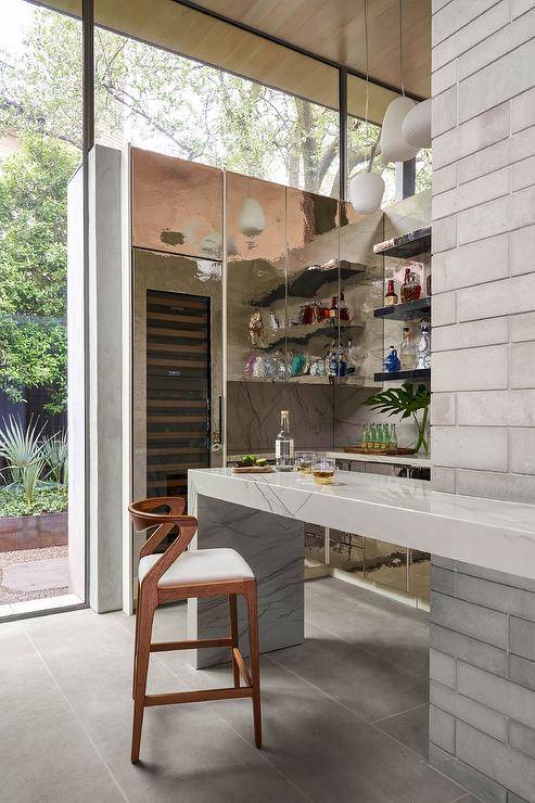 Stylish wet bar boasts a tall glass front wine fridge fixed beneath and beside iridescent flat front cabinets mounted against a marble slab backsplash, adjacent to stacked black shelves. A wooden bar stool is placed at a marble waterfall edge island.