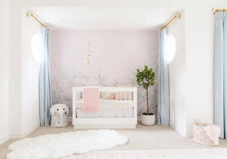 Nursery features a pink floral mural on accent wall with a gold mobile sits over a white wooden nursery crib with animal print bedding, baby blue curtains, a white wicker elephant hamper and a large white faux fur rug.