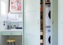 A stacked white front loading washer and dryer is hidden in floor-to-ceiling mint-green cabinets. Beside the cabinets, a hairpin stool sits on black and white cement floor tiles at a mint-green built-in desk fixed beneath stacked white shelves.