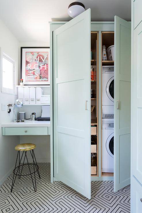 A stacked white front loading washer and dryer is hidden in floor-to-ceiling mint-green cabinets. Beside the cabinets, a hairpin stool sits on black and white cement floor tiles at a mint-green built-in desk fixed beneath stacked white shelves.