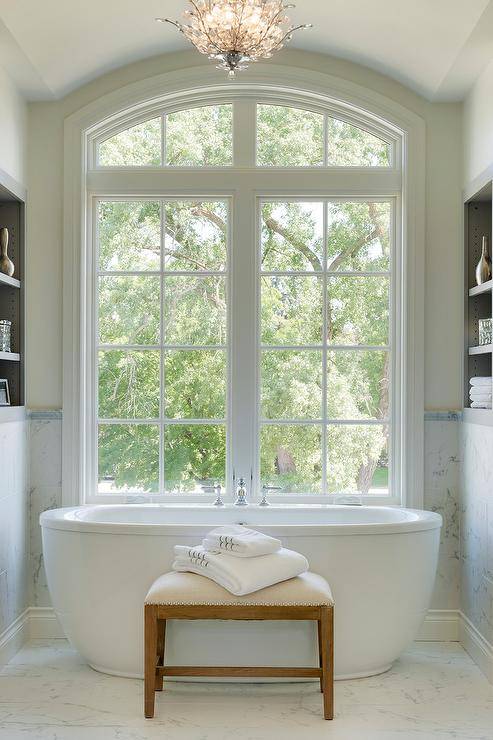 Freestanding oval tub under arched windows in a transitional master bathroom boasting a bathtub bench atop marble floor tiles.