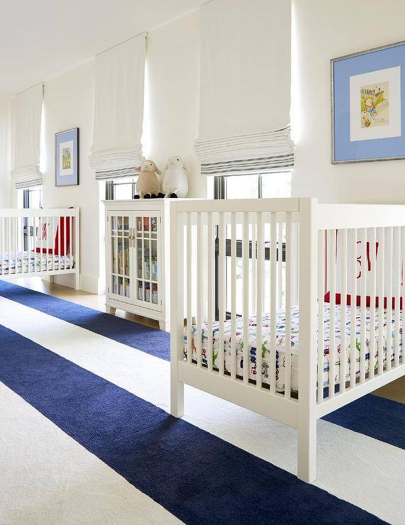 Spacious twin nursery design features a navy blue awning stripe rug, white wooden nursery cribs and a white glass front cabinet.