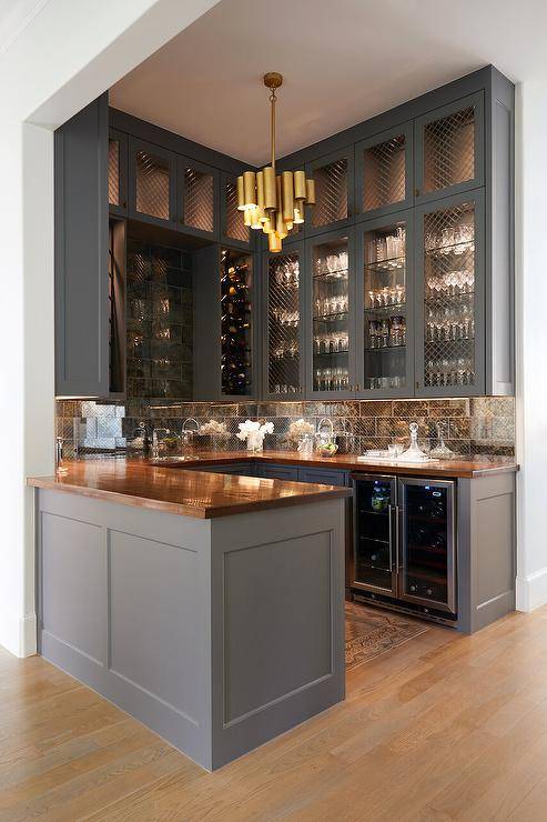 Charcoal gray u-shaped wet bar boasts charcoal gray cabinets accented with a glossy wooden countertop fitted over a glass front wine cooler. Stacked upper cabinets finished with brass grill doors are lighted and mounted against antique mirrored subway tiles.