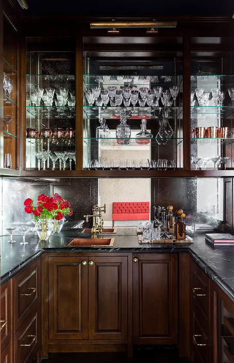 Gorgeous u-shaped wet bar boasts brown cabinets adorned with brass hardware and a beveled honed black marble countertop fixed against a mirrored backsplash. A copper hammered overmount sink matched with an antique brass wing arm faucet mounted under glass shelves.