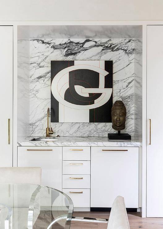 White flat-front wet bar cabinets accented with polished brass bulls and a marble countertop are built into a dining room alcove. The cabinets are finished with a sink and an aged brass gooseneck faucet mounted in front of a marble slab backsplash complemented with a black and white abstract art piece.