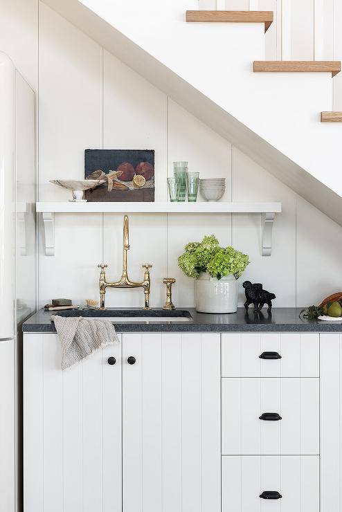 Oil rubbed bronze hardware and a black marble countertop accents white plank cabinets fixed beneath a staircase and fitted with a sink and a polished nickel deck mount faucet. A white shelf is mounted to white shiplap trim over the sink.
