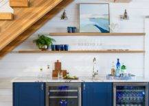 A blue wet bar under a staircase features a marble countertop with blue cabinets, stacked floating shelves and a glass front mini beverage fridge.