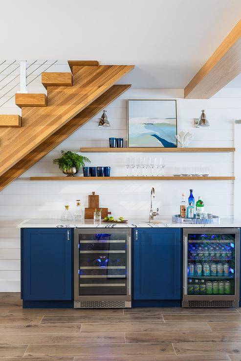 A blue wet bar under a staircase features a marble countertop with blue cabinets, stacked floating shelves and a glass front mini beverage fridge.