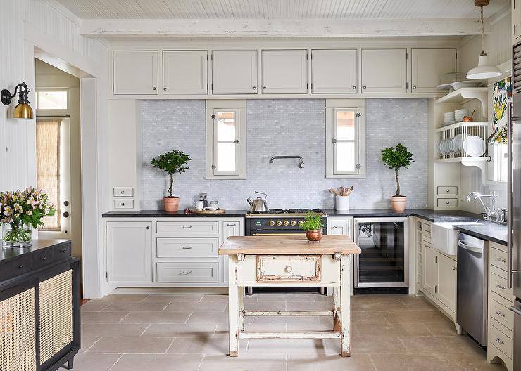Cottage kitchen design features an ivory painted reclaimed wood island, white cabinets and large beige floor tiles.