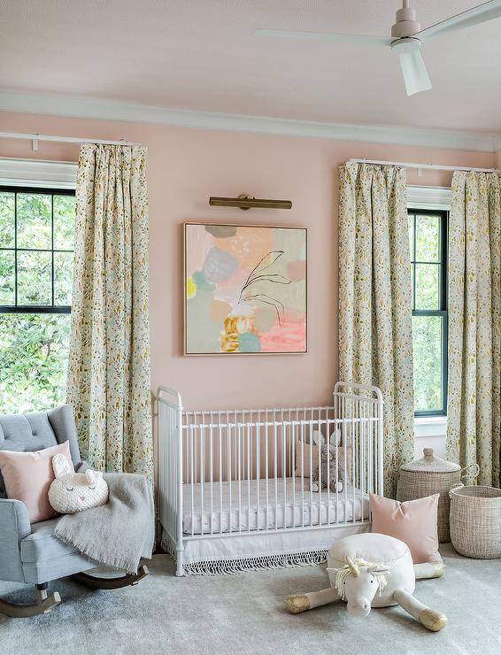Beautifully designed girl's nursery features a white metal vintage crib accented with a tassel skirt and placed on a gray rug beneath a pastel abstract art piece hung from a pink wall lit by a brass picture light. A gray tufted wingback rocker is topped with a pink pillow.
