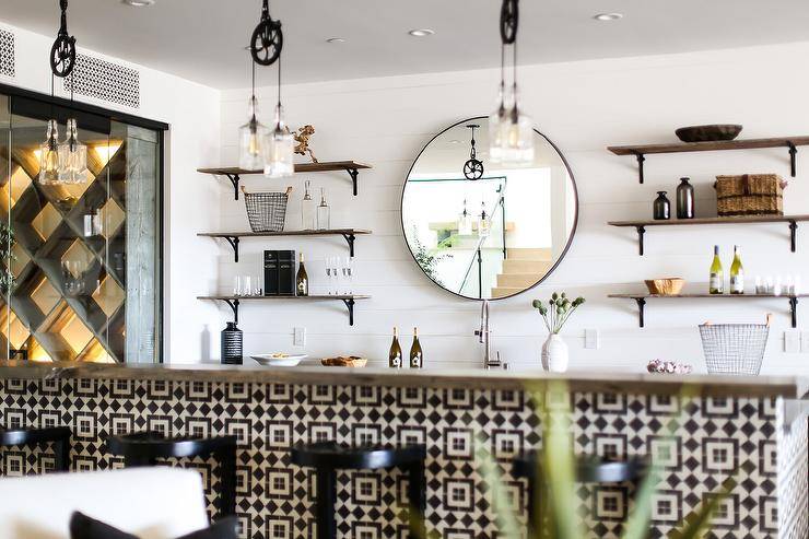 Stacked wood and metal shelves are mounted in a wet bar against a shiplap wall and on either side of a round black mirror hung over a sink. Black backless stools sit at a white and black tiled island lit by vintage staggered lanterns.