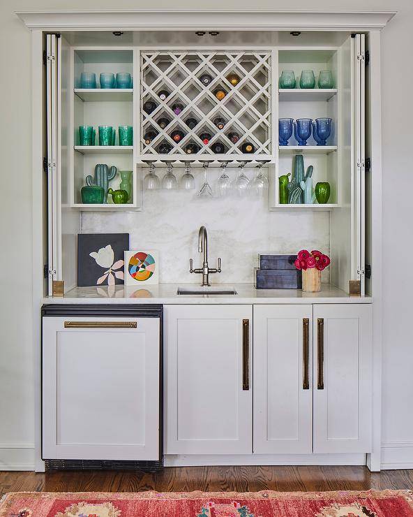 Folding doors conceal a wet bar nook boasting white shaker cabinets accented with long brass hardware and a honed marble countertop holding a small sink under a brushed nickel faucet. The faucet is fixed in front of a honed marble backsplash and under a wine rack flanked by white shelves.