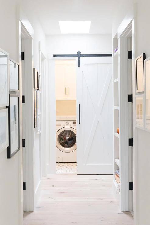 A small white laundry room is concealed behind a white barn door on rails. White shaker cabinets are mounted over a white front-loading washer and dryer.