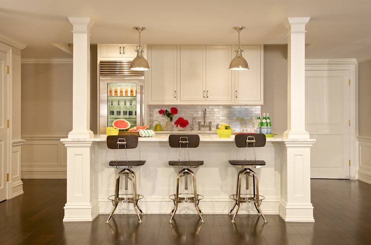 Basement kitchenette features white shaker cabinets paired with white marble countertops and stainless steel brick tile backsplash. Kitchen cabinets are suspended over sink with bridge, gooseneck faucet situated next to glass-door beverage refrigerator. Polished nickel, industrial pendants illuminate kitchen island lined with Restoration Hardware Vintage Toledo Bar Chairs Polished Chrome.