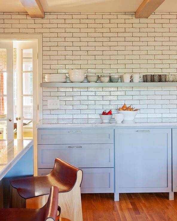 A light blue floating shelf is mounted to white offset backsplash tiles over light blue cabinets donning polished nickel pulls.