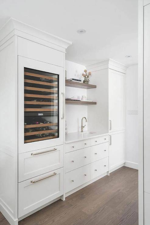 White kitchen wet bar boasts white drawers donning polished nickel ball knobs and a small sink with a polished nickel gooseneck faucet fixed beneath stacked brown wood floating shelves. The shelves are mounted between a glass front wine cooler and white cabinets.