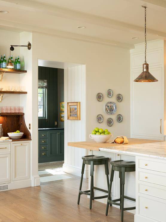 Two backless black wooden stools sit at a white center island accented with cabinets donning small brass ball knobs and topped with a mixed wood and marble countertop lit by vintage copper lanterns.