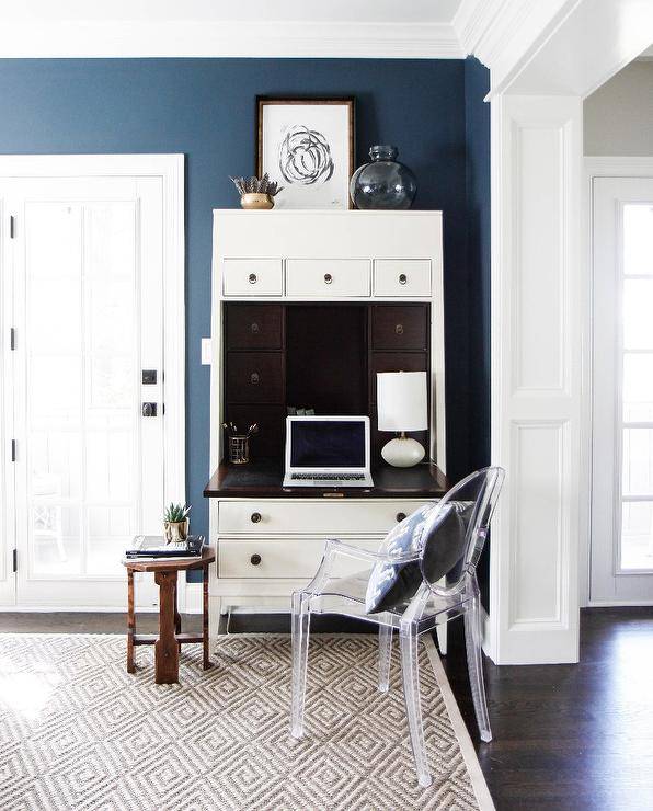 Living room office space with a Secretary desk accented with a ghost Chair topped with a blue and taupe accent pillow and sat on a diamond print jute rug also holding a small wood hexagon side table. On top of the desk a framed painting leans against blue walls complimented by a blue vase positioned beside the painting.