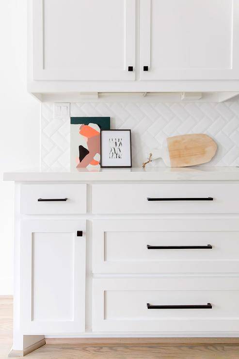 Gorgeous white kitchen boasts white shaker drawers adorned with oil rubbed bronze pulls and a complemented with white herringbone backsplash tiles installed vertically.