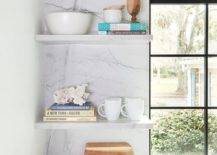 A mini brass picture light fixed to a marble slab backsplash lights stacked thin marble floating shelves positioned beside a window.