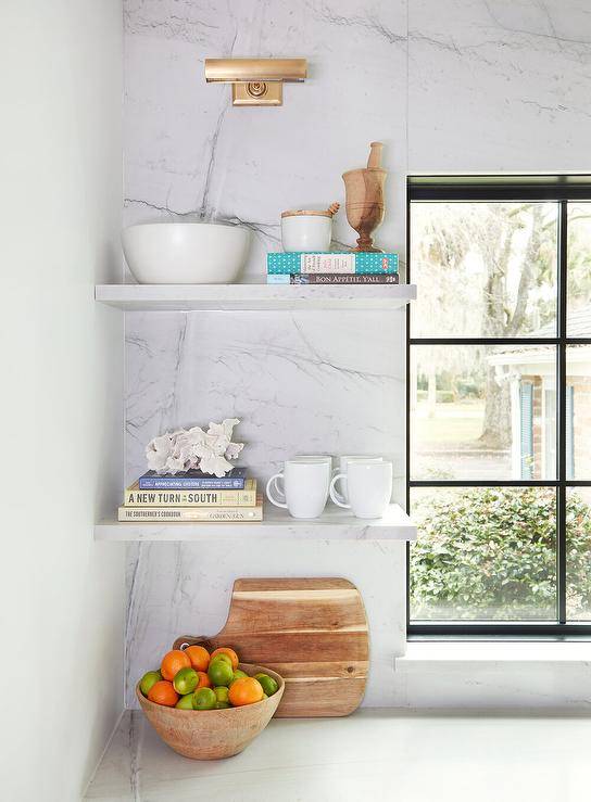 A mini brass picture light fixed to a marble slab backsplash lights stacked thin marble floating shelves positioned beside a window.