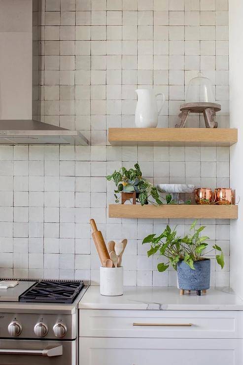 Stacked oak floating shelves are mounted against gray glazed grid tiles beside a stainless steel range hood mounted over a stainless steel oven range. White kitchen cabinets are topped with a white quartz countertop.