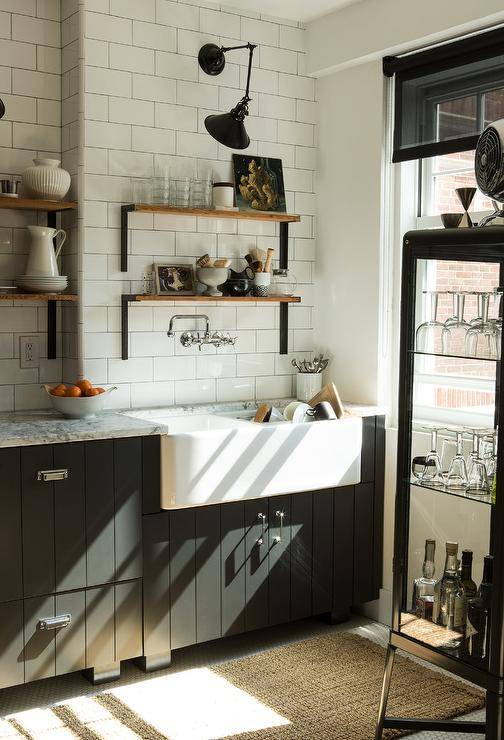 This two-toned, black and white vintage kitchen features a jute runner placed in front of black plank cabinets accented with polished nickel hardware and fitted with dual farmhouse sink matched with a polished nickel wall mount faucet. Above the faucet, wood and metal shelves are mounted to white subway backsplash tiles.