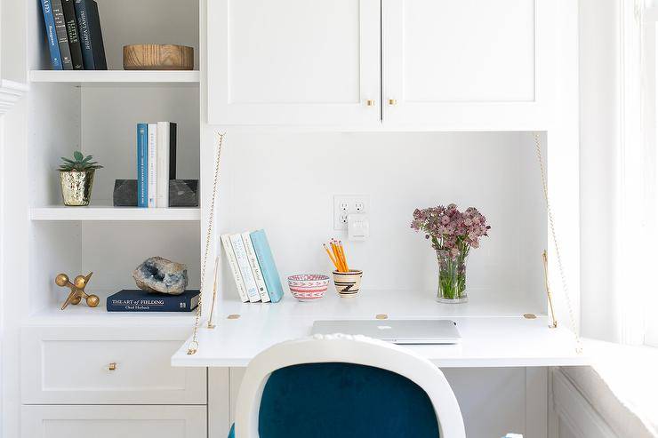 A blue velvet French Louis chair sits in front of a white pull down desk fixed under white cabinets with brass knobs and beside styled open built in cabinets in a gorgeous white living room decorated with blue accents.