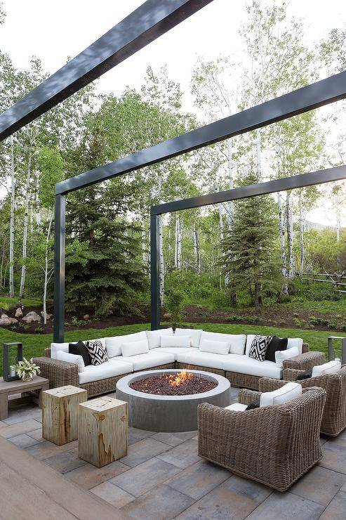 Backyard patio features a brown wicker outdoor sectional with white cushions, a round concrete fire pit, and brown wicker chairs under a modern black aluminum pergola.