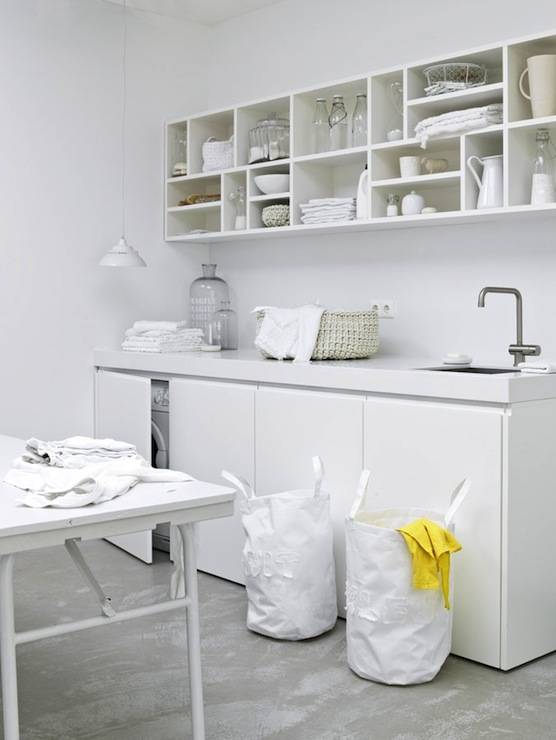 Laundry room features open display cabinets over concealed washer and dryer hidden behind cabinets paired with thick white quartz countertops and small square sink accented with satin nickel faucet. White laundry room with white folding laundry table and white hampers over concrete floor.