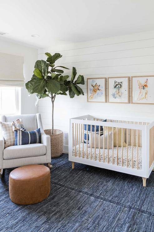 A fiddle leaf fig sits on a blue jute rug in a stunning nursery boasting a white rocker topped with blue and beige pillows and paired with a round brown leather pouf. Baby animal art hangs from shiplap trim over a two-tone vintage style crib.