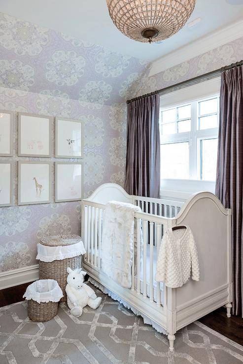 White and purple nursery features a white French wily nursery crib on a gray trellis rug, an art gallery on purple medallion wallpaper, woven baskets and purple silk curtains.