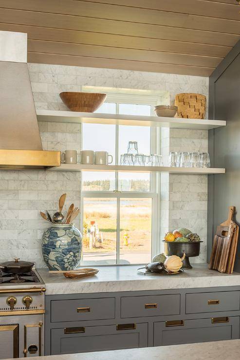 Cottage style kitchen features stacked shelves in front of a kitchen window with marble offset backsplash tiles and gray cabinets adorned with vintage brass hardware.