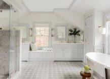 Luxurious master bathroom features a marble hexagon floor tiles leading towards a a light gray built-in window seat topped with a white cushion. The windows seat is flanked by light gray his and hers washstands.