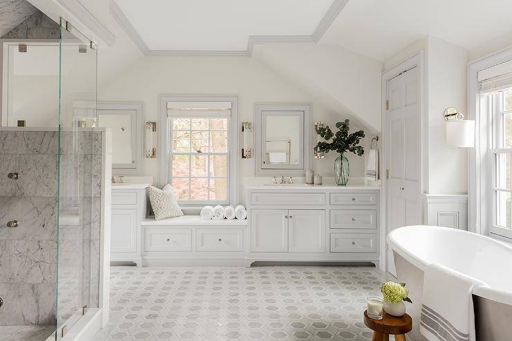 Luxurious master bathroom features a marble hexagon floor tiles leading towards a a light gray built-in window seat topped with a white cushion. The windows seat is flanked by light gray his and hers washstands.